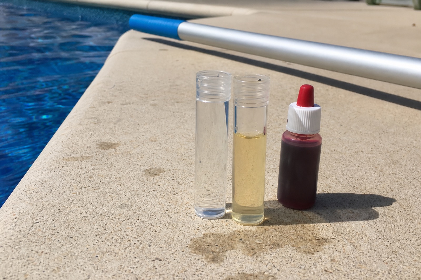 Pool water test vials on the deck — one clear, one tinted from phenol red — beside a small reagent bottle.